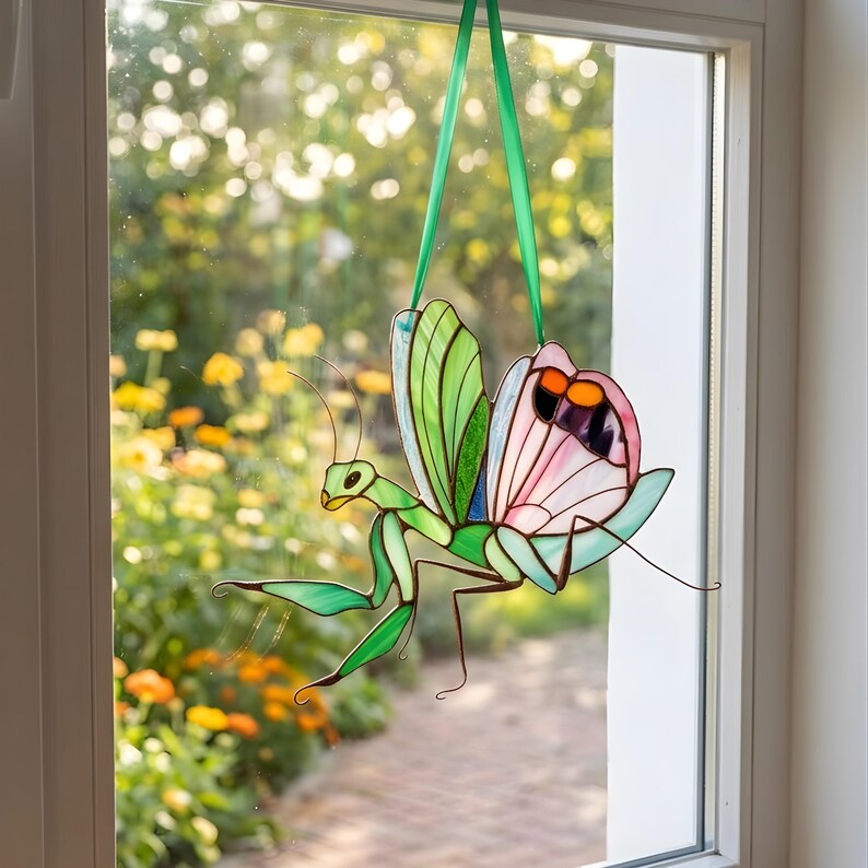 Stained glass praying mantis suncatcher hanging on window, sunlight shining through colorful glass wings, handmade insect decor creating soft light reflections.