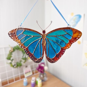 May include: A stained glass butterfly with blue, teal, orange, and red wings. The butterfly is hanging from a blue ribbon.
