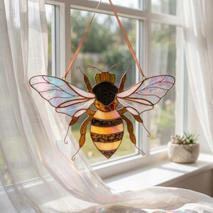 Stained glass bee suncatcher hanging in window with soft curtain and sunlight. Iridescent wings shimmer with rainbow colors while amber glass body glows warmly, creating bright window decor.