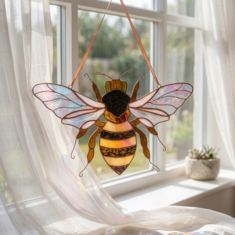 Stained glass bee suncatcher hanging in window with soft curtain and sunlight. Iridescent wings shimmer with rainbow colors while amber glass body glows warmly, creating bright window decor.