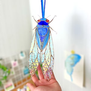 May include: A stained glass cicada with iridescent wings. The cicada is blue and yellow with red eyes. The wings are made of small pieces of glass that are arranged in a pattern. The cicada is hanging from a blue ribbon.