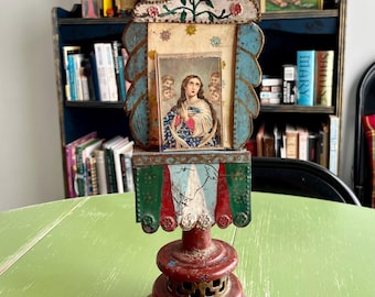 May include: A vintage tin religious icon with a colorful painted frame and a picture of the Virgin Mary. The icon is on a red metal base with a decorative design.