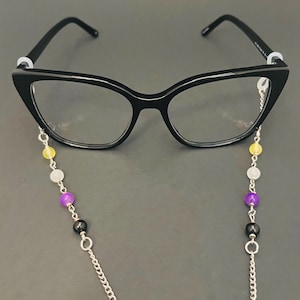 May include: Black cat-eye glasses with a beaded eyeglass chain. The chain features a silver chain with colorful beads in yellow, white, purple, and black. The glasses are on a dark gray background.