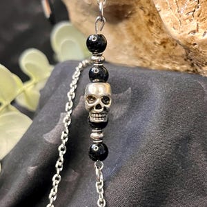 May include: A silver-toned necklace featuring a skull pendant and black beads. The skull is detailed with visible teeth and eye sockets. The chain is made of small, interconnected links. The beads are round and smooth, adding contrast to the skull.