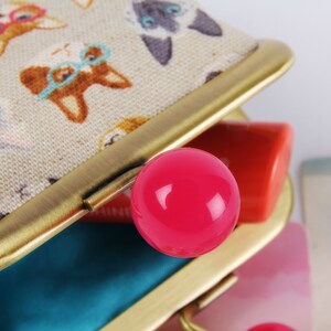 May include: A small, beige fabric coin purse with a gold metal frame and a pink plastic button. The purse is open and has a blue lining. The fabric has a pattern of cats wearing glasses.