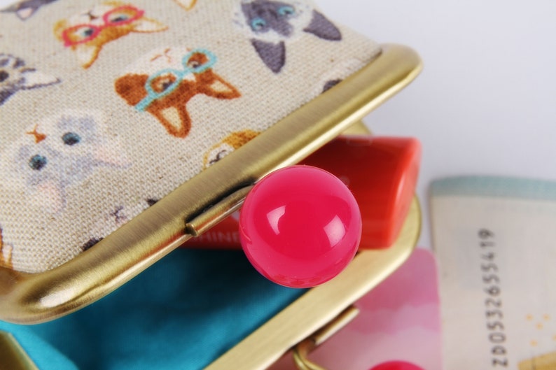 May include: A small, beige fabric coin purse with a gold metal frame and a pink plastic button. The purse is open and has a blue lining. The fabric has a pattern of cats wearing glasses.