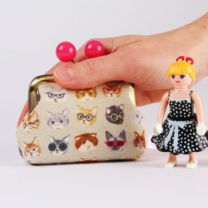 May include: A small beige coin purse with a gold clasp and pink buttons. The purse is covered in a pattern of cats wearing glasses. A small doll with blonde hair and a black and white polka dot dress is standing next to the purse.