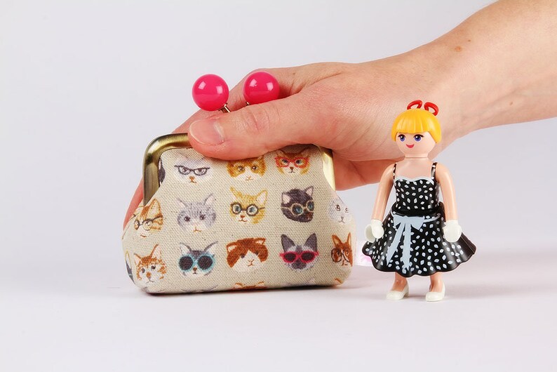 May include: A small beige coin purse with a gold clasp and pink buttons. The purse is covered in a pattern of cats wearing glasses. A small doll with blonde hair and a black and white polka dot dress is standing next to the purse.