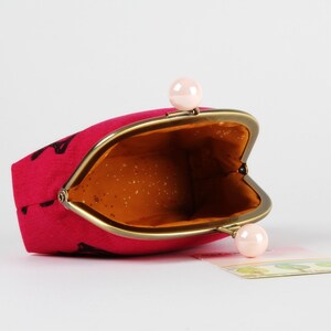 Op de afbeelding: Een roze stoffen muntportemonnee met een gouden metalen frame en twee roze parel-achtige sluitingen. De portemonnee is open en de binnenkant is gevoerd met een bruine stof.