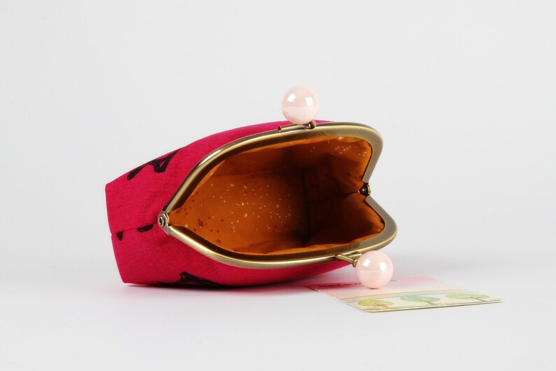 Op de afbeelding: Een roze stoffen muntportemonnee met een gouden metalen frame en twee roze parel-achtige sluitingen. De portemonnee is open en de binnenkant is gevoerd met een bruine stof.