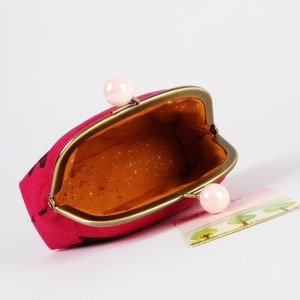 Op de afbeelding: Een roze stoffen muntportemonnee met een gouden metalen frame en twee roze plastic sluitingen. De portemonnee is open en de binnenkant is gevoerd met een bruine stof.
