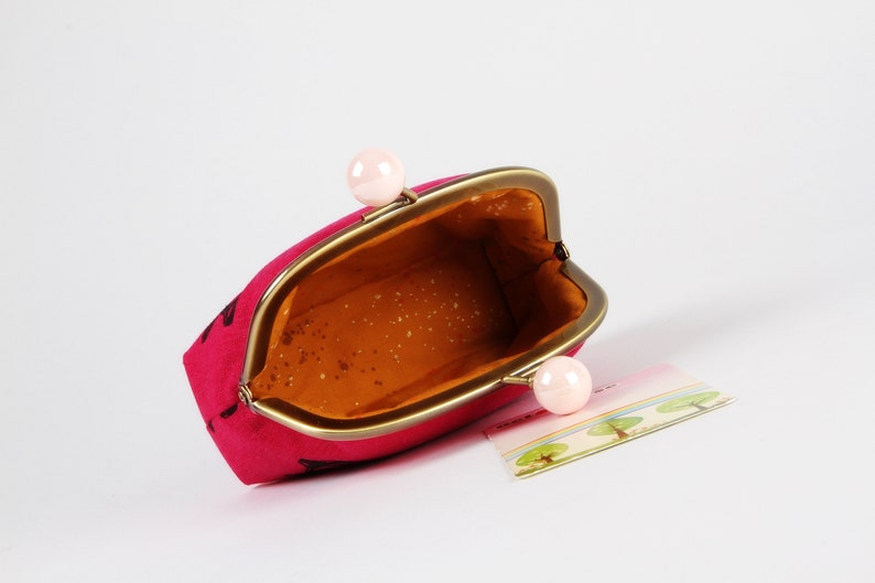 Op de afbeelding: Een roze stoffen muntportemonnee met een gouden metalen frame en twee roze plastic sluitingen. De portemonnee is open en de binnenkant is gevoerd met een bruine stof.