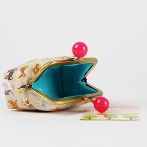 May include: A small, beige fabric coin purse with a gold metal frame and pink plastic closures. The purse is open and the inside is lined with turquoise fabric.