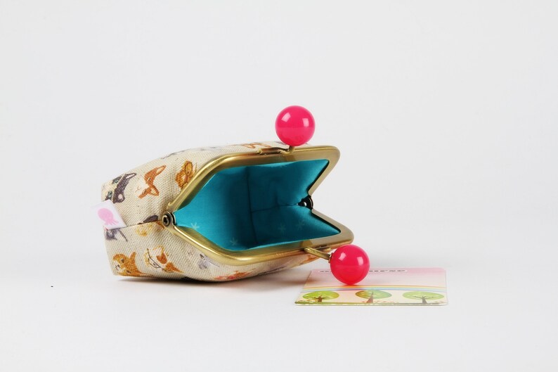 May include: A small, beige fabric coin purse with a gold metal frame and pink plastic closures. The purse is open and the inside is lined with turquoise fabric.