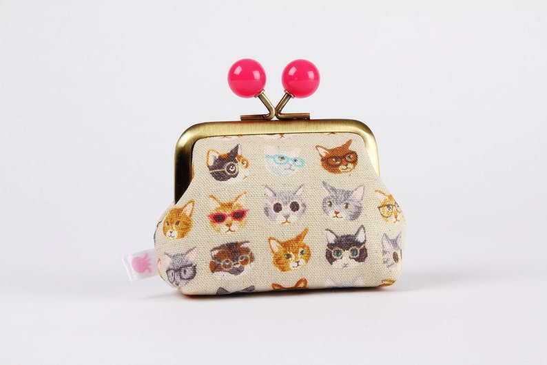 May include: A small, beige coin purse with a gold metal frame and pink plastic clasps. The purse is covered in a pattern of cats wearing glasses.