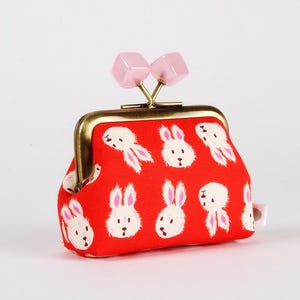 Peut inclure: Un porte-monnaie rouge avec un fermoir en laiton et des accents de cubes roses. Le sac est orné de lapins blancs de dessins animés. Un accessoire mignon pour transporter de petits objets.