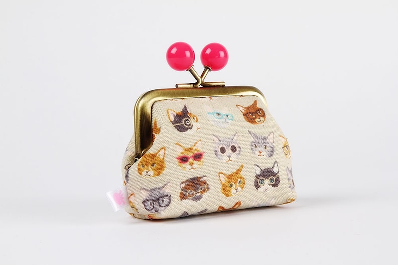 May include: A small beige fabric coin purse with a gold metal frame and pink plastic clasps. The purse is decorated with a repeating pattern of cats wearing glasses.