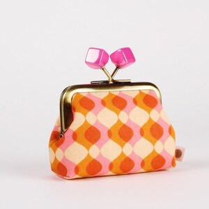 May include: A small coin purse with a retro design and a kiss-lock clasp. The purse has a repeating pattern of orange, pink, and cream-coloured curved shapes. The clasp is gold-toned, and the top is decorated with two pink square accents.