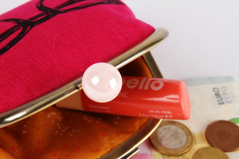 Op de afbeelding: Een roze stoffen muntportemonnee met een gouden metalen frame en een roze knoop. De portemonnee is open en er zit een tube lippenbalsem in met het woord "bello" erop en wat euro-munten.