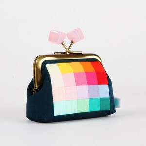 Peut inclure: Un porte-monnaie bleu canard avec un motif patchwork coloré sur le devant. Le sac à main a un fermoir doré et deux accents roses en forme de carré sur le dessus. Le patchwork présente des carrés de différentes couleurs.
