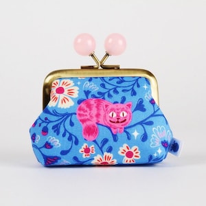Pode incluir: Uma bolsa azul com um padrão floral e um design de gato rosa. A bolsa tem uma moldura dourada e fechos de bola rosa. O tecido apresenta flores brancas e rosas.