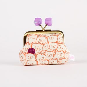 May include: A small, cream-colored coin purse with a repeating pattern of orange cat faces. The purse has a gold-colored metal frame and a clasp with two purple square accents. A small ball of yarn is attached to the front.