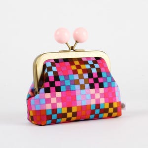 Peut inclure: Un porte-monnaie coloré avec un motif pixelisé rose, bleu, marron et noir. Il est doté d'un fermoir doré et de deux ornements roses en forme de boule. Une petite étiquette blanche est attachée sur le côté.