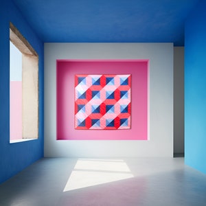 May include: A geometric quilt with red, blue and pink squares arranged in a pattern. The quilt is hanging on a wall in a room with blue and pink walls.