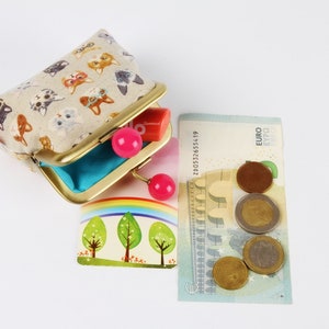 May include: A small, beige fabric coin purse with a gold metal frame and pink plastic clasps. The purse is decorated with a pattern of cats. The purse is open and a small, pink plastic lipstick is visible inside. A euro bill and several euro coins are visible in the background.