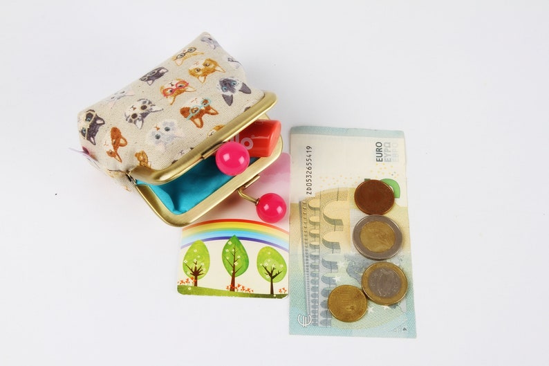May include: A small, beige fabric coin purse with a gold metal frame and pink plastic clasps. The purse is decorated with a pattern of cats. The purse is open and a small, pink plastic lipstick is visible inside. A euro bill and several euro coins are visible in the background.