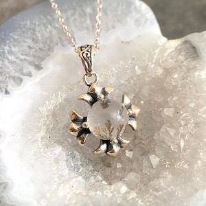 May include: A silver pendant necklace with a clear quartz crystal featuring dendritic inclusions. The pendant is shaped like a four-pointed star with ornate details.