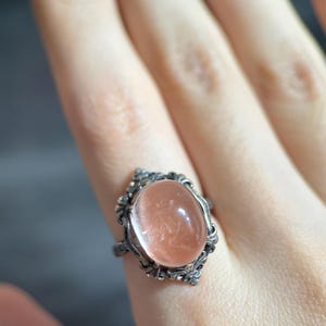 Op de afbeelding: Een zilverkleurige ring met een ovale, perzikroze edelsteen. De steen is gezet in een decoratief, sierlijk montuur. De ring wordt aan een vinger gedragen, waarbij de ingewikkelde details van het sieraad worden getoond.