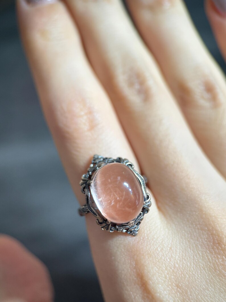 Op de afbeelding: Een zilverkleurige ring met een ovale, perzikroze edelsteen. De steen is gezet in een decoratief, sierlijk montuur. De ring wordt aan een vinger gedragen, waarbij de ingewikkelde details van het sieraad worden getoond.