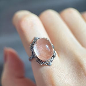 Op de afbeelding: Een zilveren ring met een ovale, doorschijnende, perzik-kleurige edelsteen. De ring heeft een sierlijk filigraanontwerp rond de steen en op de band. De ring wordt aan een vinger getoond.
