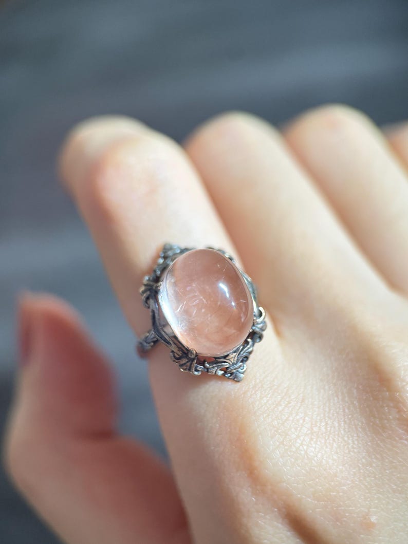 Op de afbeelding: Een zilveren ring met een ovale, doorschijnende, perzik-kleurige edelsteen. De ring heeft een sierlijk filigraanontwerp rond de steen en op de band. De ring wordt aan een vinger getoond.