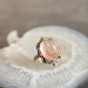 Op de afbeelding: Een ring in vintage-stijl met een grote, ovale, perzik-kleurige edelsteen. De ring heeft een sierlijke, gedetailleerde zetting met een zilverkleurige band en goudkleurige accenten. De edelsteen is glad en doorschijnend.