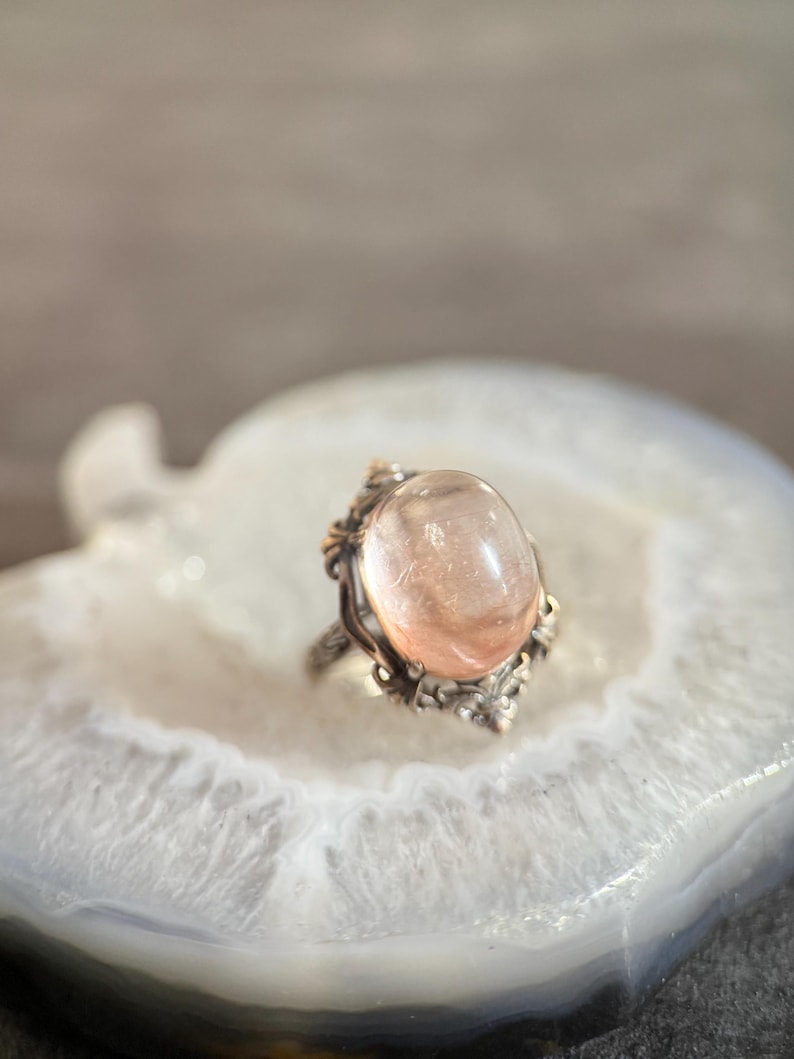 Op de afbeelding: Een ring in vintage-stijl met een grote, ovale, perzik-kleurige edelsteen. De ring heeft een sierlijke, gedetailleerde zetting met een zilverkleurige band en goudkleurige accenten. De edelsteen is glad en doorschijnend.