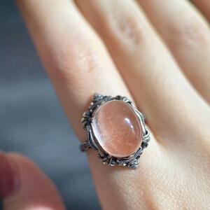 Op de afbeelding: Een zilveren ring met een ovale, perzik-kleurige edelsteen. De ring heeft een sierlijk filigraan ontwerp rond de steen. De ring zit aan een vinger.