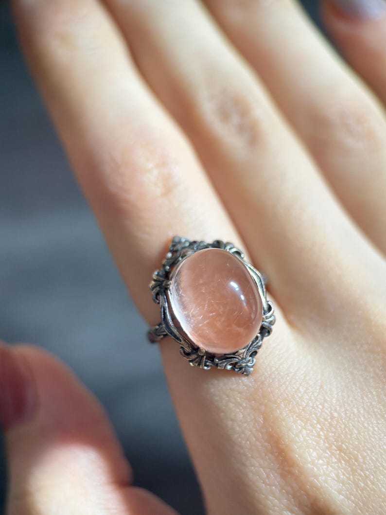 Op de afbeelding: Een zilveren ring met een ovale, perzik-kleurige edelsteen. De ring heeft een sierlijk filigraan ontwerp rond de steen. De ring zit aan een vinger.