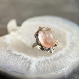 Op de afbeelding: Een zilveren ring met een grote, ovale, perzik-kleurige edelsteen. De ring heeft sierlijke details rond de steen en is gezet op een witte en grijze agaatsteen. Het ontwerp van de ring bevat gouden accenten.