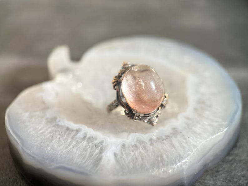 Op de afbeelding: Een zilveren ring met een grote, ovale, perzik-kleurige edelsteen. De ring heeft sierlijke details rond de steen en is gezet op een witte en grijze agaatsteen. Het ontwerp van de ring bevat gouden accenten.