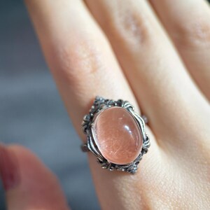 Op de afbeelding: Een zilveren ring met een ovale, perzik-kleurige edelsteen. De edelsteen is gezet in een decoratief, sierlijk frame. De ring wordt op een vinger getoond, waarbij het ontwerp en de kleur van het sieraad worden benadrukt.