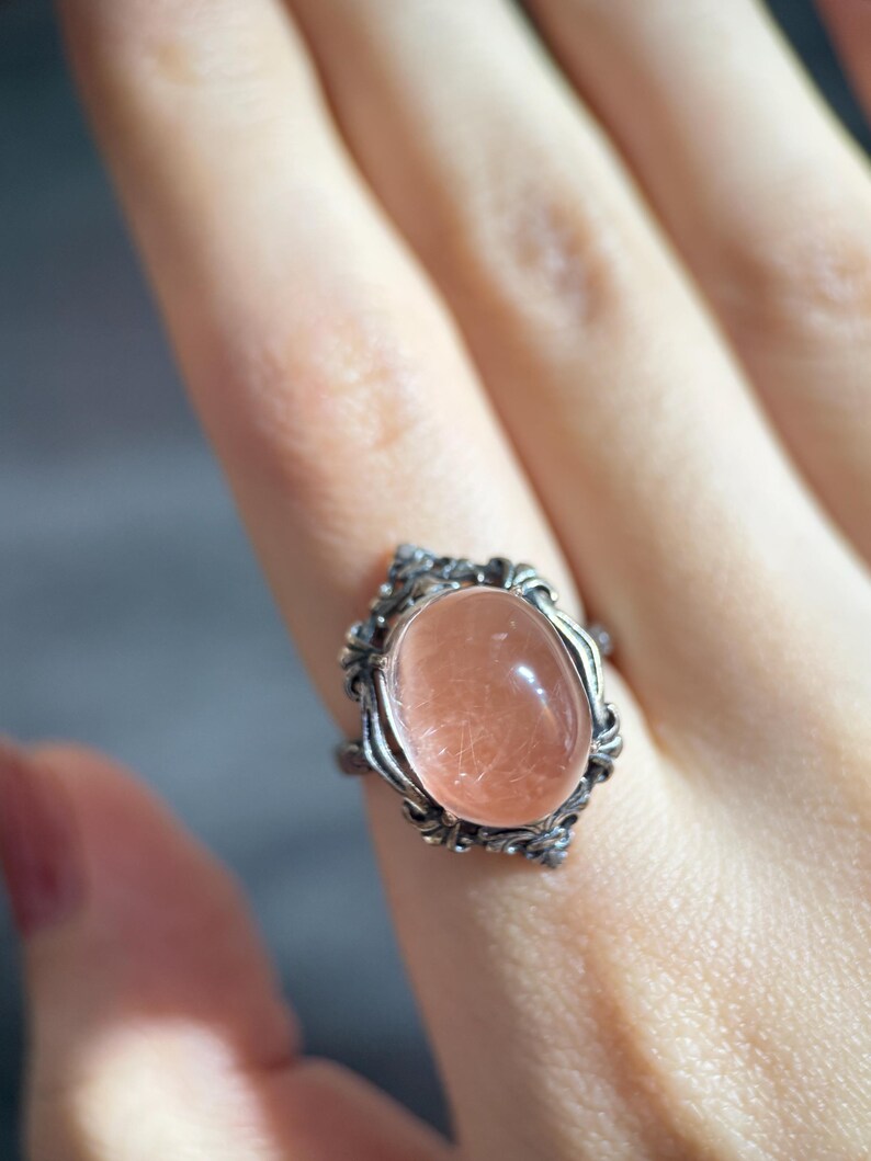 Op de afbeelding: Een zilveren ring met een ovale, perzik-kleurige edelsteen. De edelsteen is gezet in een decoratief, sierlijk frame. De ring wordt op een vinger getoond, waarbij het ontwerp en de kleur van het sieraad worden benadrukt.
