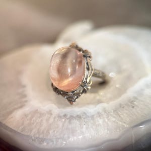 Op de afbeelding: Een zilveren ring met een ovale, perzik-kleurige edelsteen. De ring heeft een sierlijke zetting met bloemen- en krulmotieven. De edelsteen is geplaatst op een wit, agaatachtig oppervlak.