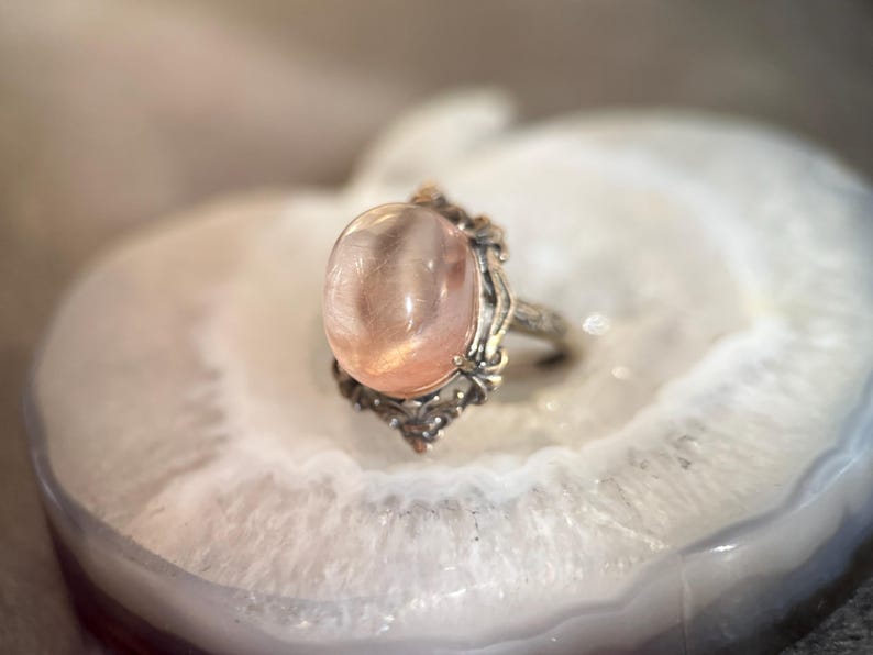 Op de afbeelding: Een zilveren ring met een ovale, perzik-kleurige edelsteen. De ring heeft een sierlijke zetting met bloemen- en krulmotieven. De edelsteen is geplaatst op een wit, agaatachtig oppervlak.