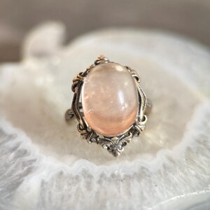 Op de afbeelding: Een vintage ring met een ovale, lichtroze edelsteen, gezet in een sierlijk, zilverkleurig metalen frame. Het ontwerp van de ring heeft ingewikkelde filigraan details rond de edelsteen, wat een klassieke en elegante uitstraling cre&euml;ert. De ring wordt gepresenteerd op een wit, gemarmerd oppervlak.
