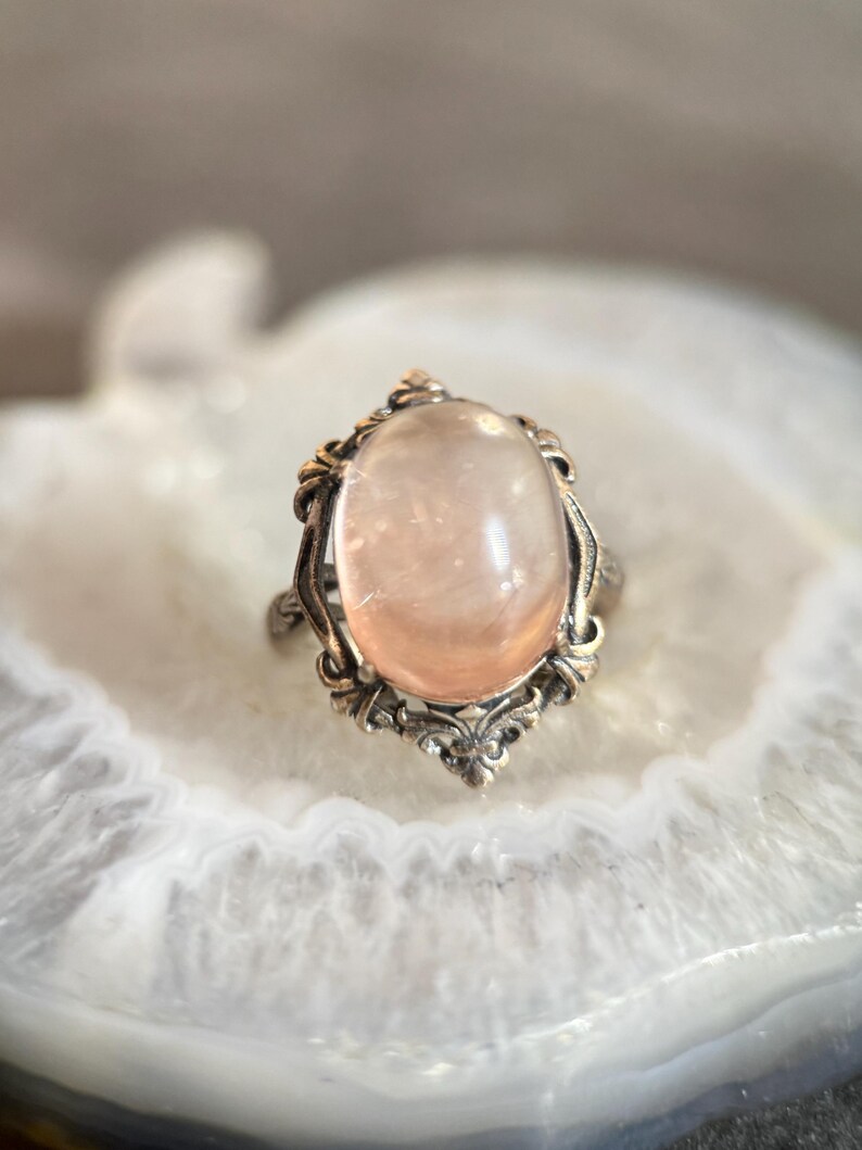 Op de afbeelding: Een vintage ring met een ovale, lichtroze edelsteen, gezet in een sierlijk, zilverkleurig metalen frame. Het ontwerp van de ring heeft ingewikkelde filigraan details rond de edelsteen, wat een klassieke en elegante uitstraling cre&euml;ert. De ring wordt gepresenteerd op een wit, gemarmerd oppervlak.