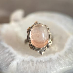 Op de afbeelding: Een zilveren ring met een grote, ovale, perzik-kleurige edelsteen. De ring heeft een sierlijke, vintage-stijl zetting met gouden accenten. De doorschijnende edelsteen heeft zichtbare interne structuren. De ring wordt gepresenteerd op een wit en grijs oppervlak.