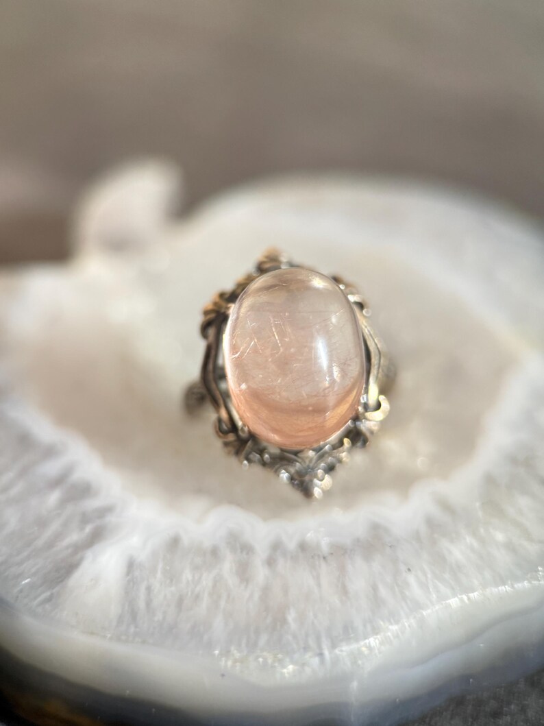 Op de afbeelding: Een zilveren ring met een grote, ovale, perzik-kleurige edelsteen. De ring heeft een sierlijke, vintage-stijl zetting met gouden accenten. De doorschijnende edelsteen heeft zichtbare interne structuren. De ring wordt gepresenteerd op een wit en grijs oppervlak.