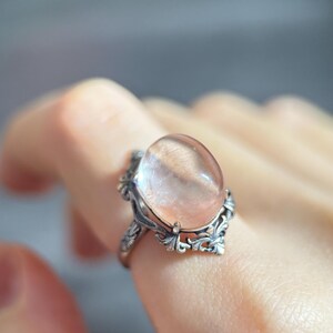 Op de afbeelding: Een zilveren ring met een ovale, doorschijnende, perzik-kleurige edelsteen. De ring heeft een sierlijk filigraan ontwerp met blad- en kruldetails rond de steen. De ring wordt aan een vinger gedragen.